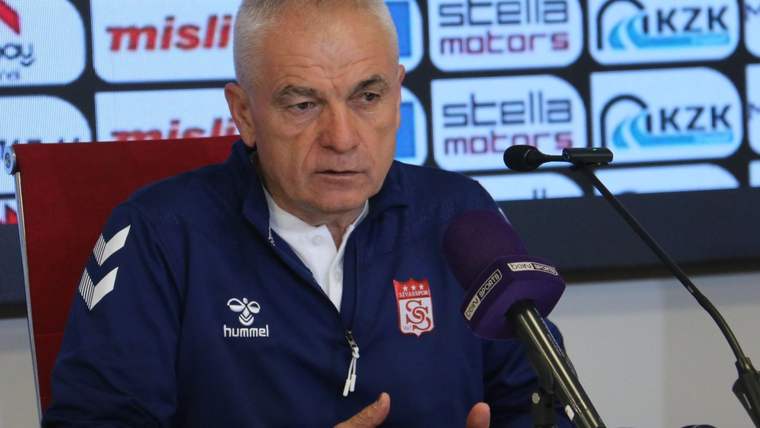Basın toplantısında, önünde mikrofonlar bulunan Sivasspor armalı eşofmanlı bir kişi konuşuyor.