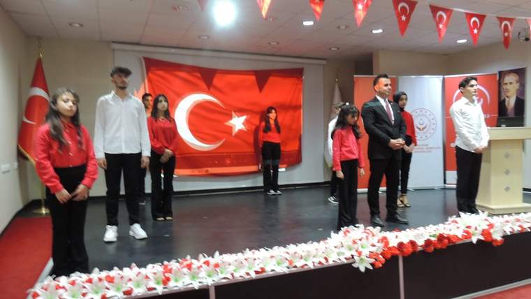 Sahne üzerinde Türk bayrağı arkasında çocuklar ve yetişkinler duruyor, bazıları beyaz, bazıları kırmızı kıyafet giymiş.
