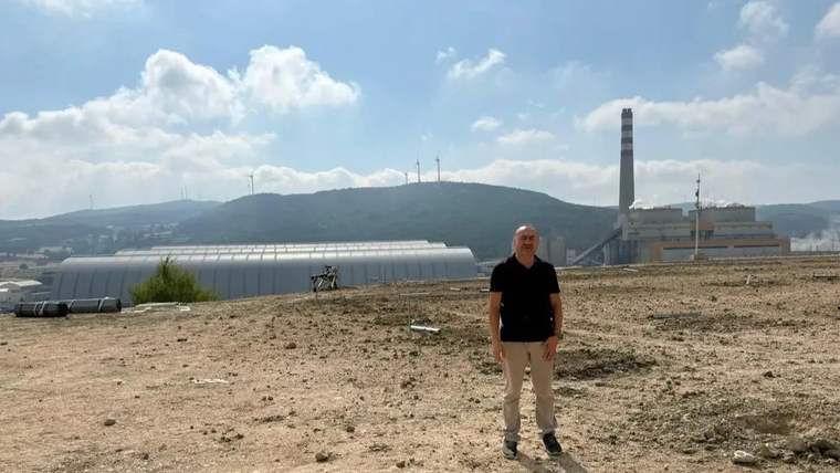 Kuru ve taşlı arazide duran bir kişi; arkasında seralar, yüksek bir baca ve tepeler görülüyor.