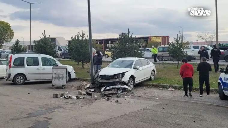 Beyaz bir araç, direğe çarparak hasar görmüş; çevresinde insanlar ve ağaçlar var.