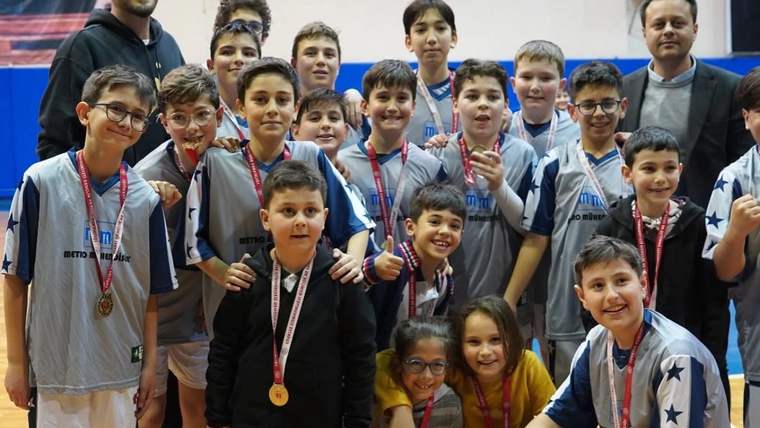 Spor salonunda madalyalı çocuk basketbolcular, iki yetişkinle birlikte kameraya poz veriyor.