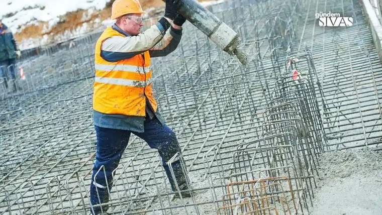 İnşaat alanında, turuncu yelek giymiş bir işçi, beton döküyor.