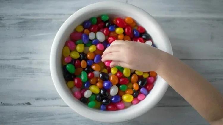 Bir çocuğun eli, renkli şekerlerle dolu beyaz bir kâsenin içinden şeker alıyor.