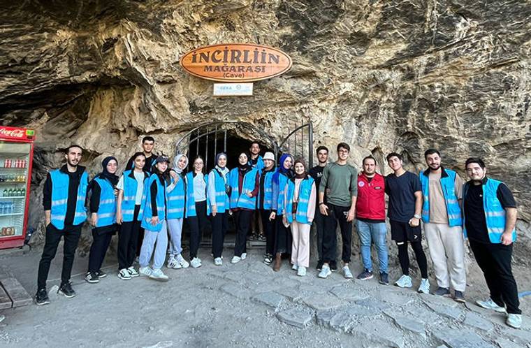 İncirliin Mağarası tabelasının önünde, mavi yelekli bir grup mağara girişinde birlikte duruyor.