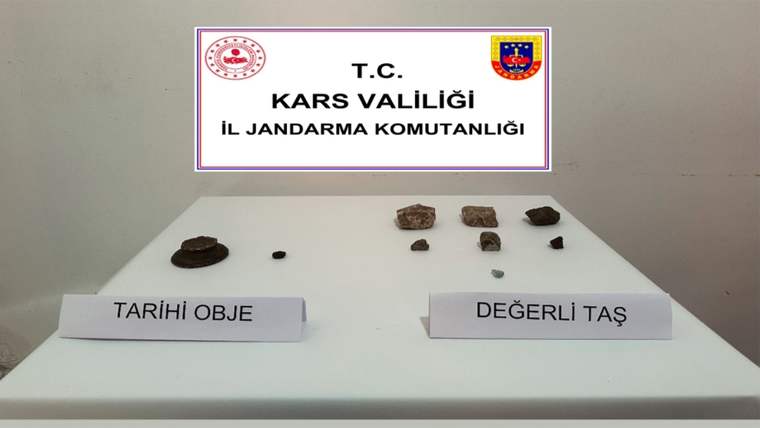 Beyaz bir masada, üzerinde 'Tarihi Obje' ve 'Değerli Taş' yazılı etiketler bulunan çeşitli taşlar yer alıyor.