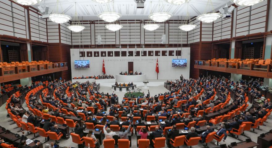 Yarım daire düzenindeki salonda çok sayıda kişi oturuyor; kürsünün arkasında iki Türk bayrağı ve ekranlar bulunuyor.