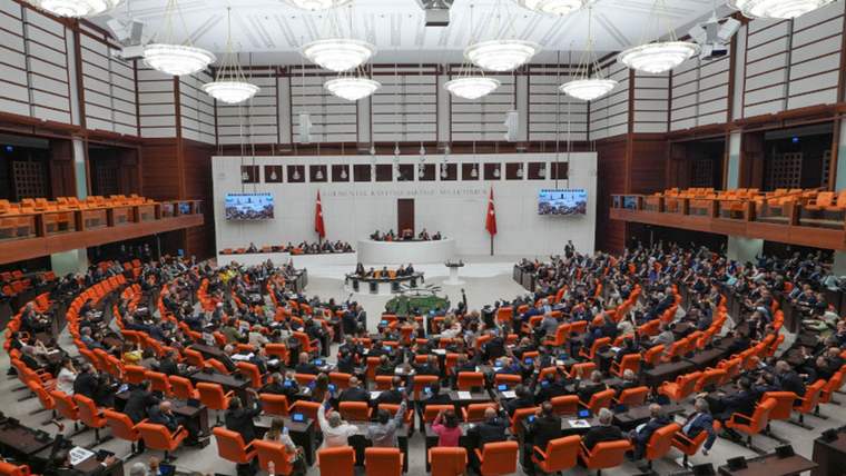 Yarım daire düzenindeki salonda çok sayıda kişi oturuyor; kürsünün arkasında iki Türk bayrağı ve ekranlar bulunuyor.