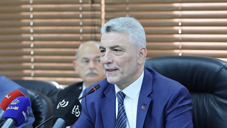 Takım elbiseli bir kişi, mikrofonların önünde masada otururken konuşuyor; arkada iki kişi görülüyor.