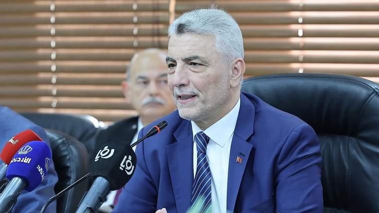 Takım elbiseli bir kişi, mikrofonların önünde masada otururken konuşuyor; arkada iki kişi görülüyor.
