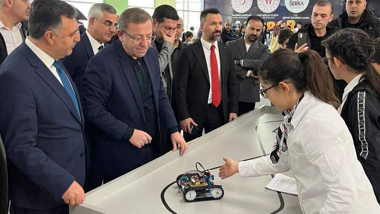 Bir grup yetişkin, masadaki küçük paletli robotu incelerken beyaz önlüklü iki öğrenci yanlarında duruyor.
