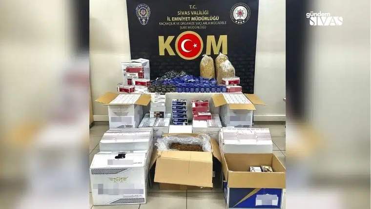 KOM yazılı pano önünde, kutular ve tezgâh üzerinde çok sayıda sigara paketi ile tütün ürünleri sergileniyor.