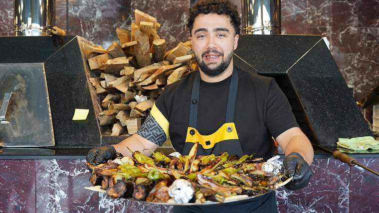 Ellerinde kebap ve sebzelerle dolu büyük bir tepsi tutan bir kişi, arka planda odunlar görünmekte.