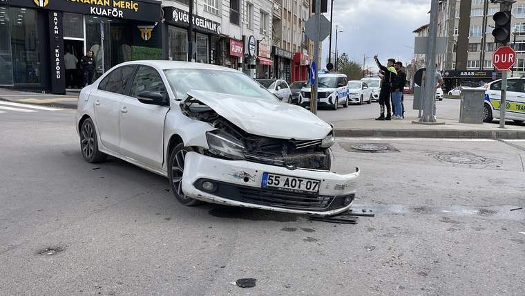 Ön kısmı ağır hasar gören beyaz otomobil kavşakta duruyor; arka planda polis araçları ve iş yerleri bulunuyor.