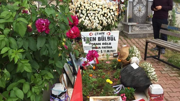 Çiçekler ve Türk bayraklarıyla süslenmiş bir mezarın yanında, telefonuna bakan bir kişi duruyor.