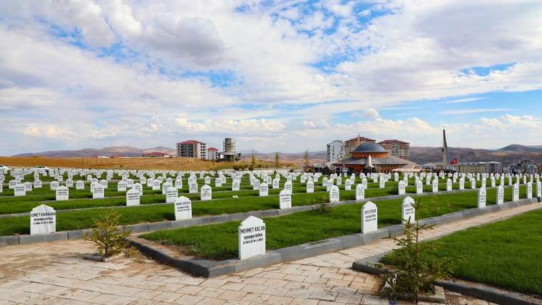 Yeşil çimlerle kaplı mezarlıkta, beyaz mezar taşları sıralanmış. Arka planda binalar ve bir cami görünmekte.