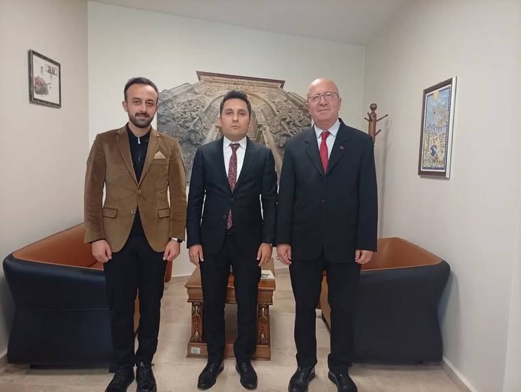 Bir odada takım elbiseli iki kişi ve kahverengi ceketli bir kişi, duvar süsünün önünde yan yana duruyor.