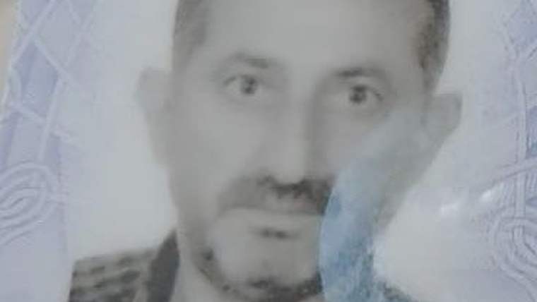 Bulanık portrede bıyıklı bir erkek, desenli bir arka plan önünde kameraya doğru bakıyor.