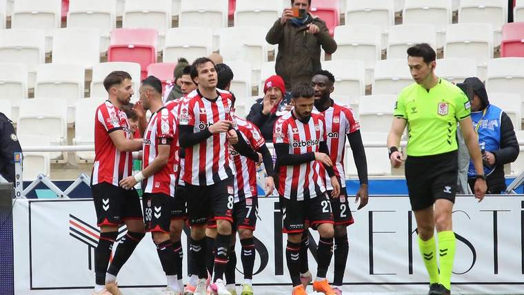 Kırmızı-beyaz çizgili formalar giyen futbolcular saha kenarında toplanırken yeşil formalı hakem yanlarından geçiyor.