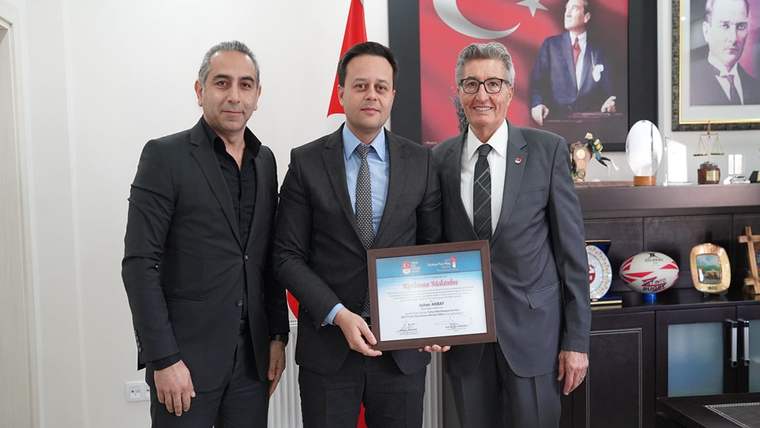 Üç erkek, bir Türk bayrağı önünde çerçeveli belge tutarak poz veriyor; arkada spor topları ve portre bulunuyor.