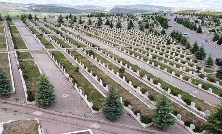 Bir mezarlık alanında, düzenli sıralanmış mezar taşları ve ağaçlar görünmektedir.