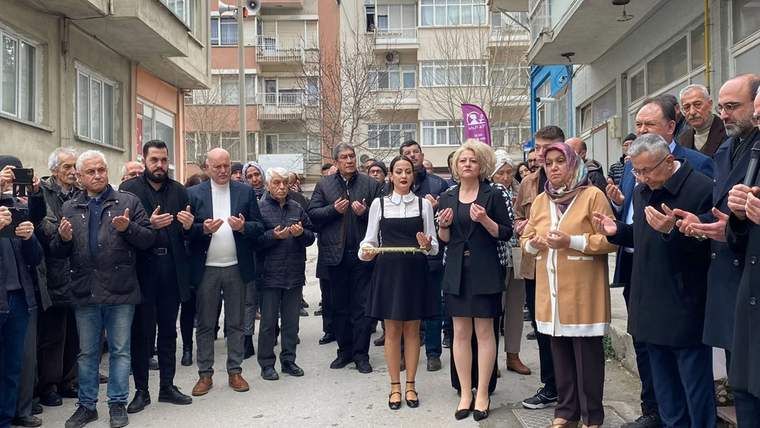 Açık alanda bir grup insan, tepsi tutan bir kadının çevresinde ellerini kaldırarak duruyor.