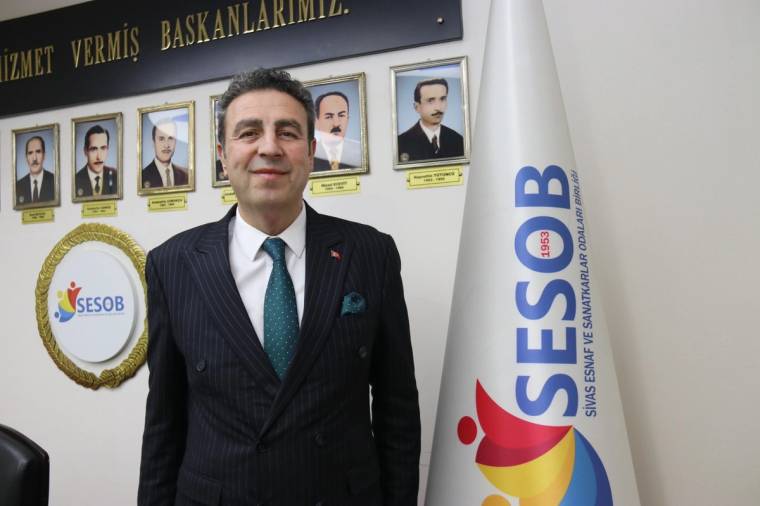 Takım elbiseli bir kişi, duvarında portreler ve SESOB yazılı bir bayrağın bulunduğu odada duruyor.