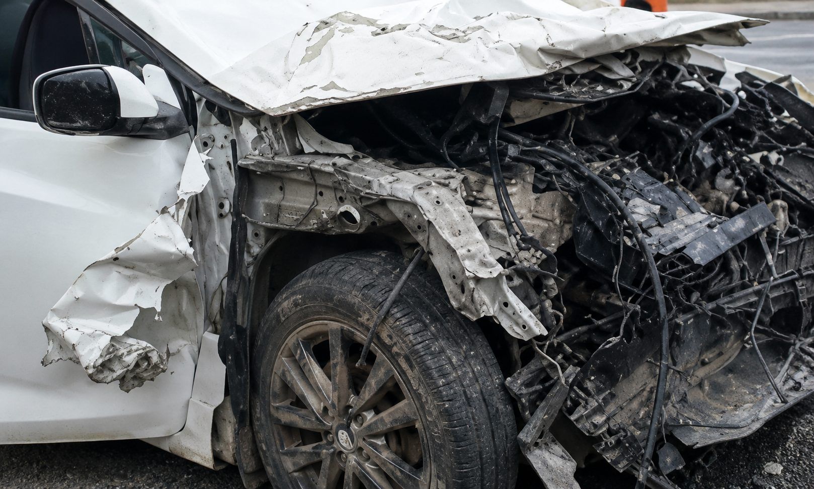 Ön kısmı ağır hasar görmüş beyaz otomobilin ezilmiş kaputu, motor bölümü ve sol ön tekerleği görülüyor.