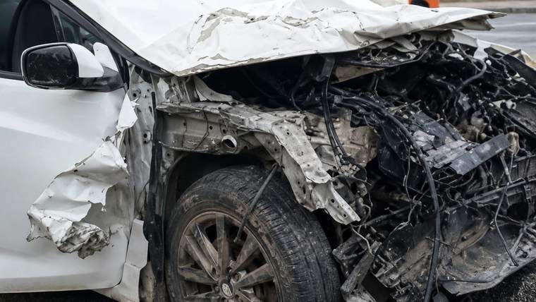 Ön kısmı ağır hasar görmüş beyaz otomobilin ezilmiş kaputu, motor bölümü ve sol ön tekerleği görülüyor.
