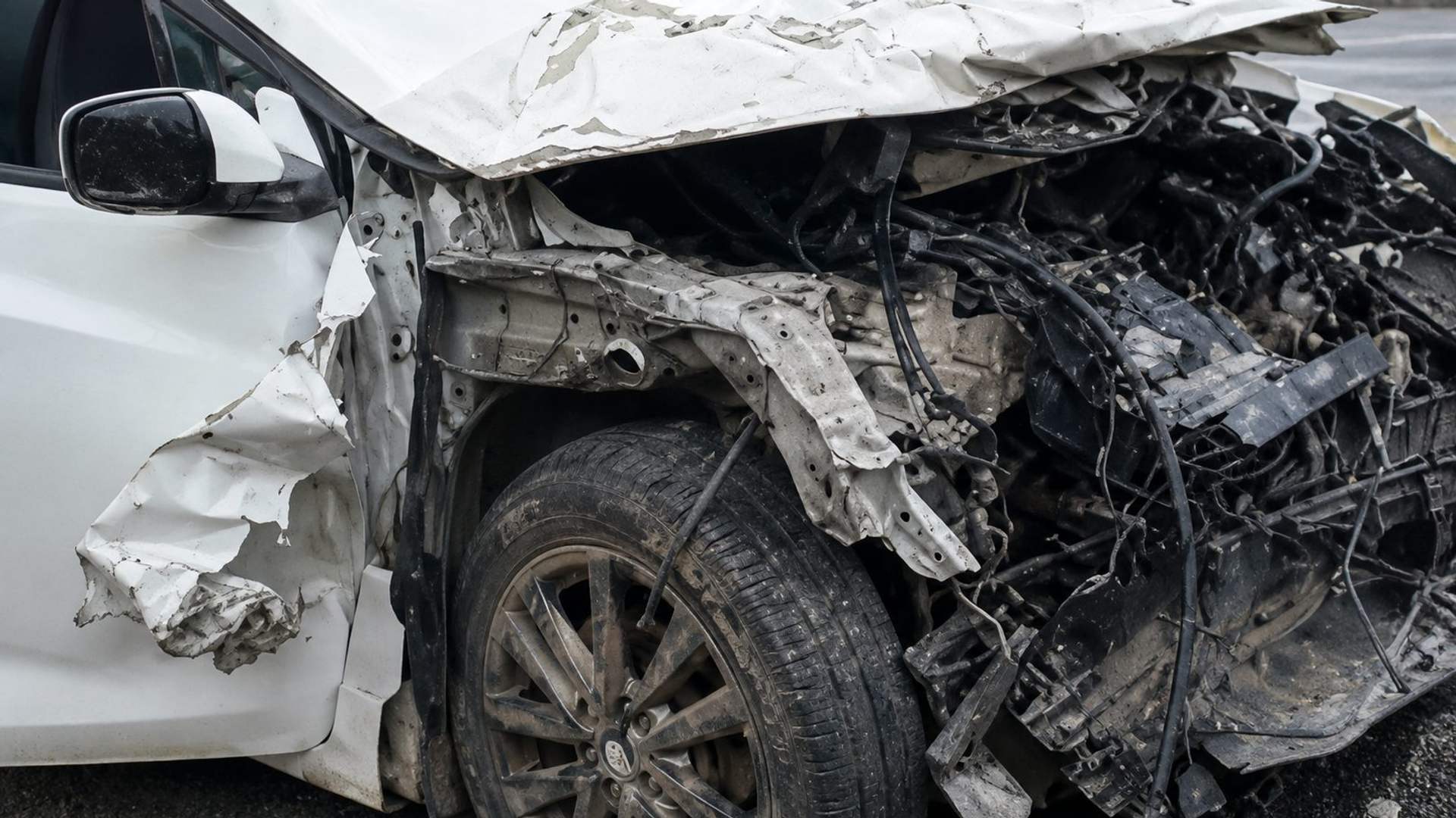 Ön kısmı ağır hasar görmüş beyaz otomobilin ezilmiş kaputu, motor bölümü ve sol ön tekerleği görülüyor.