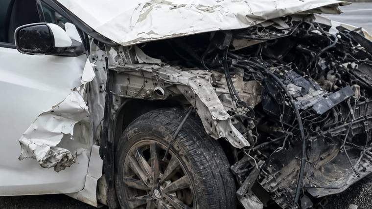Ön kısmı ağır hasar görmüş beyaz otomobilin ezilmiş kaputu, motor bölümü ve sol ön tekerleği görülüyor.