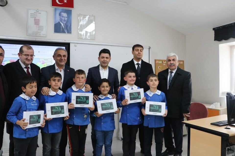 Bir grup çocuk, ellerinde tabletlerle birlikte bir sınıfta poz veriyor. Arkada birkaç yetişkin ve Türk bayrağı görünmekte.