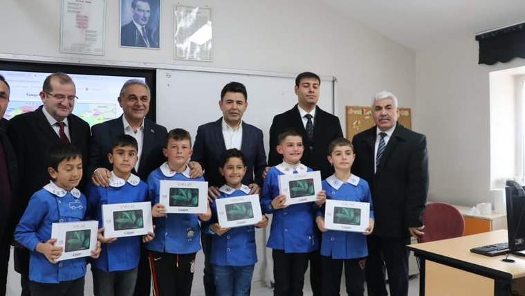 Bir grup çocuk, ellerinde tabletlerle birlikte bir sınıfta poz veriyor. Arkada birkaç yetişkin ve Türk bayrağı görünmekte.