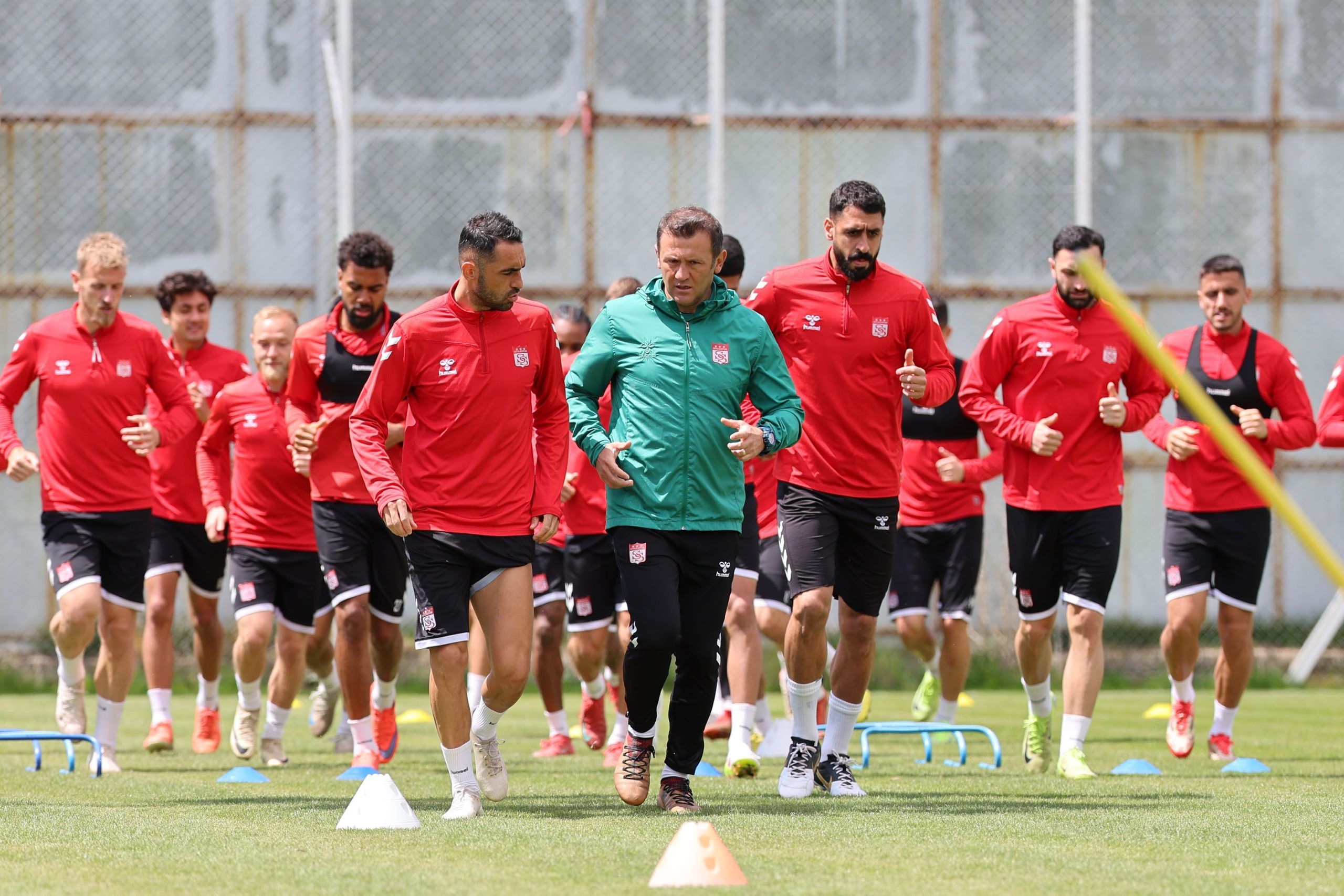 Kırmızı formalar giymiş futbolcular, antrenör eşliğinde sahada koşuyor.