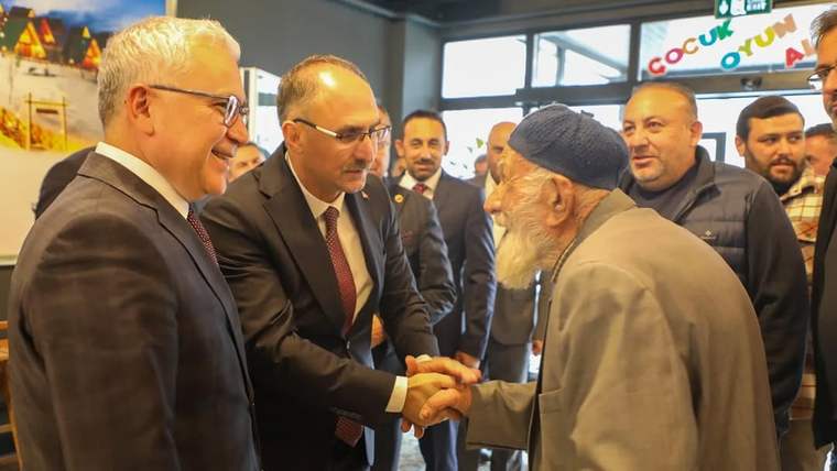 Takım elbiseli bir kişi, sakallı yaşlı bir adamla el sıkışırken çevrelerinde başka kişiler duruyor.
