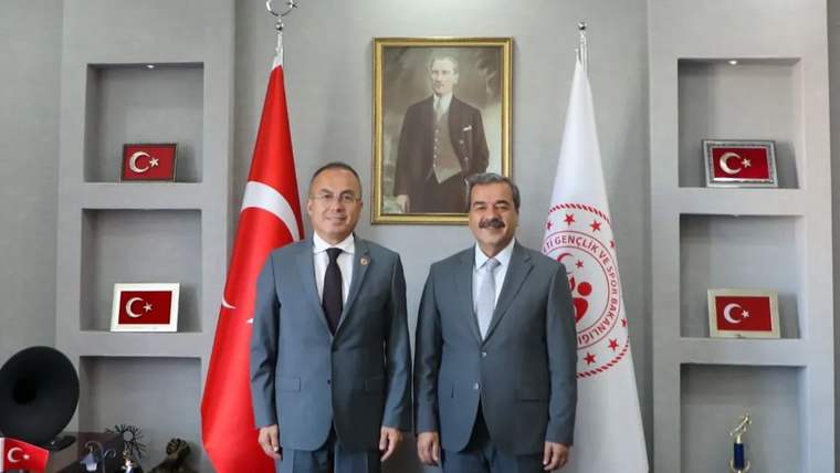 Türk bayrakları ve bir kurum flaması önünde duran takım elbiseli iki erkek, duvardaki portrenin yanında görülüyor.