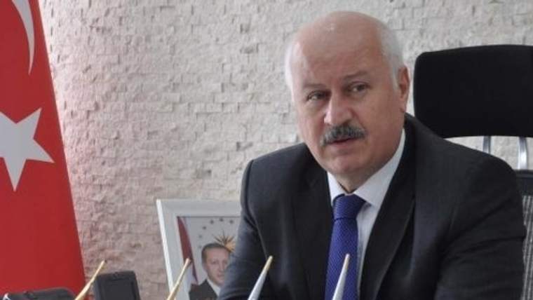 Kravatlı bir kişi, mikrofonların önünde çalışma masasında oturuyor; arka planda Türk bayrağı ve çerçeveli portre bulunuyor.