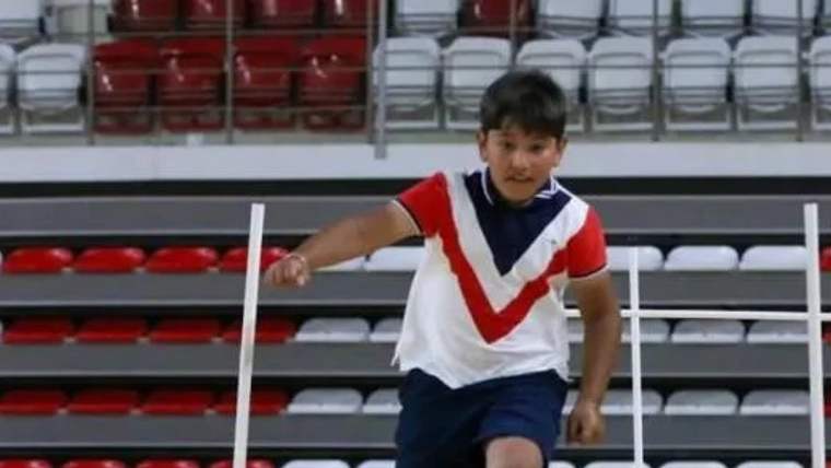 Kırmızı ve beyaz renkte tişört giymiş bir çocuk, spor salonunda engel atlama yapıyor.