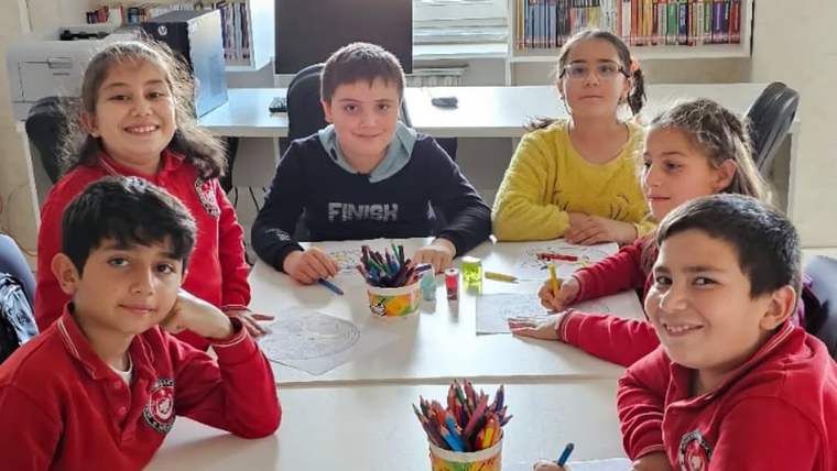 Kütüphanede bir masa çevresinde oturan altı çocuk, renkli kalemlerle kâğıt üzerinde çalışıyor.