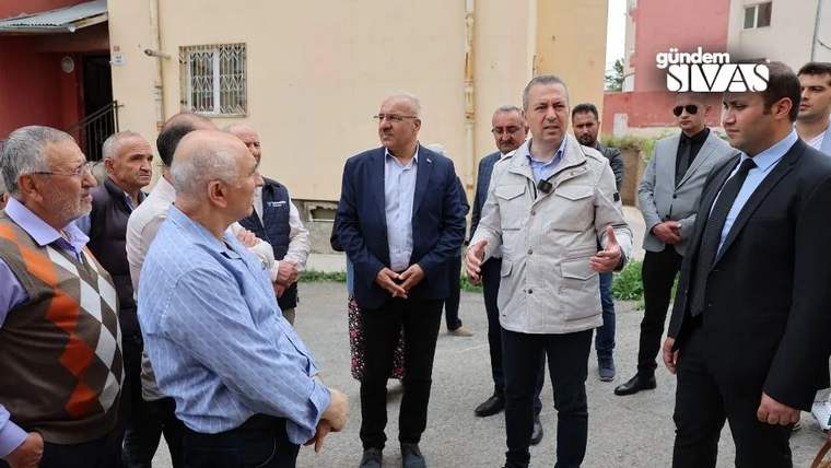 Bir grup insan, bir adamın konuşmasını dinliyor; arka planda binalar var.