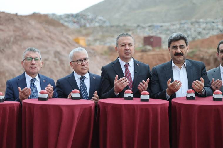 Kayalık bir alanda, kırmızı örtülü masaların arkasında duran dört kişi ellerini kaldırıyor.
