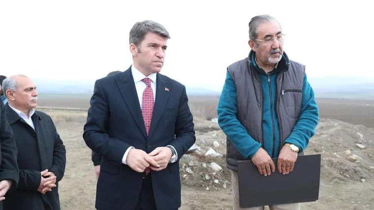 İki kişi, açık alanda durarak birbirine bakıyor; biri belgeleri tutuyor.