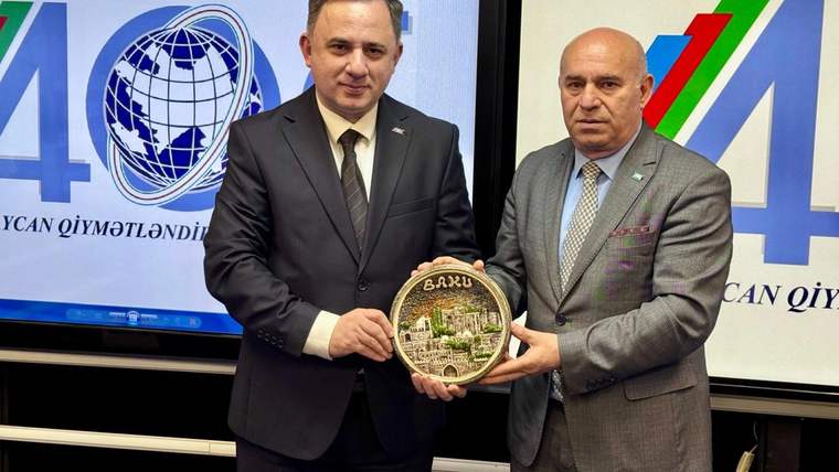 İki takım elbiseli erkek, üzerinde tarihi yapı ve “Bakı” yazan yuvarlak plaketi birlikte tutuyor.