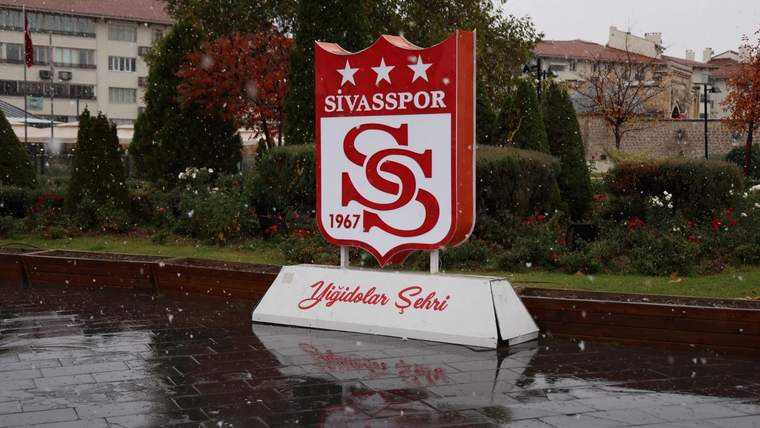 Sivasspor arması, yağış altında ıslak taş zemindeki çiçekli alanın önünde duruyor.