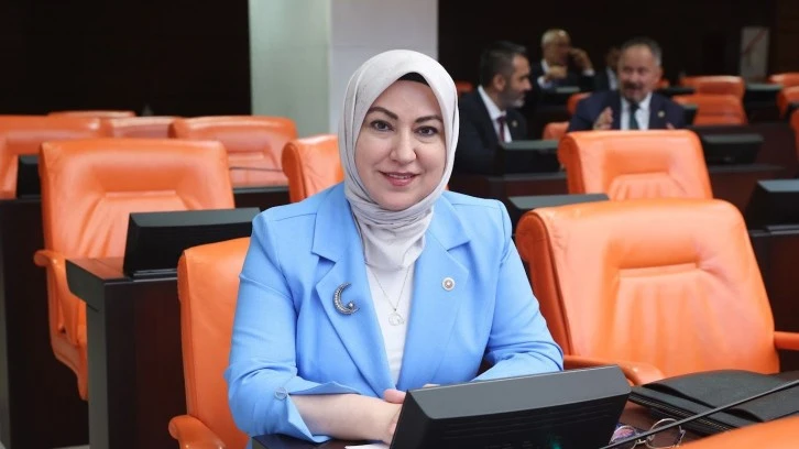 Mavi ceket ve başörtüsü takan bir kadın, oturumda oturuyor ve gülümsüyor.