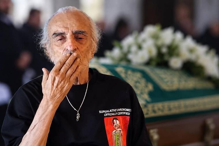 Beyaz saçlı yaşlı bir adam, arkasındaki çiçeklerle süslenmiş tabutun önünde elini ağzına götürüyor.