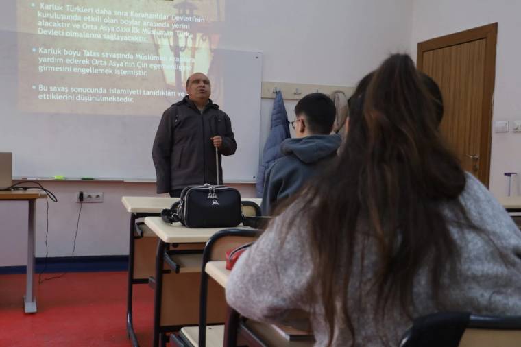 Sınıfta elinde baston tutan bir kişi, sıralarda oturan öğrencilerin karşısında konuşuyor; duvarda yansıtılmış yazılar görülüyor.