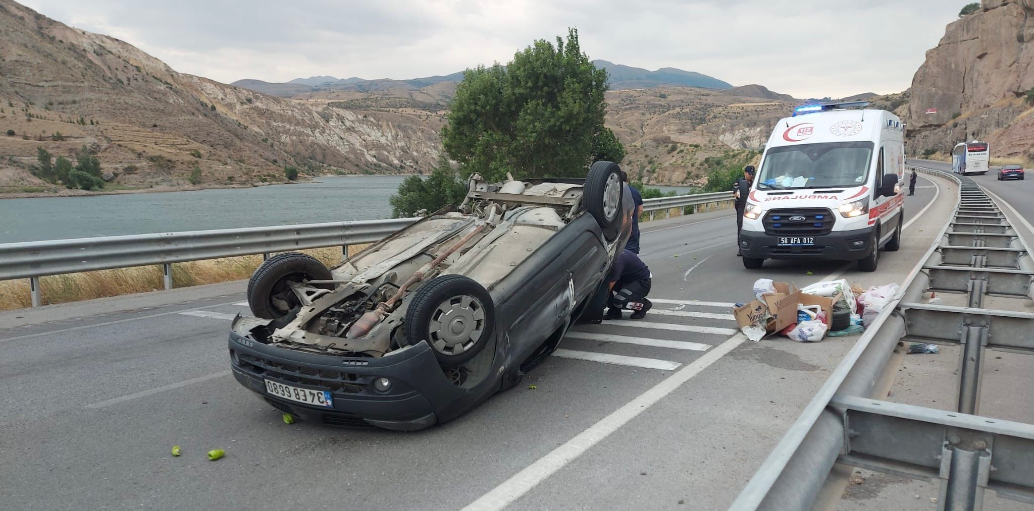 Bölünmüş yolda ters dönmüş bir otomobilin yanında ambulans ve yerde çeşitli eşyalar görülüyor.