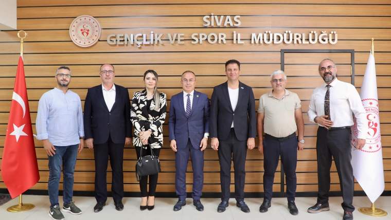 Sivas Gençlik ve Spor İl Müdürlüğü tabelası önünde, Türk bayrağı ve kurum flamaları arasında yedi kişi duruyor.