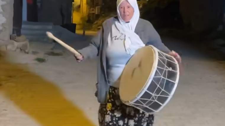 Başında beyaz örtü bulunan bir kadın, sokakta büyük bir davula tokmakla vururken görülüyor.