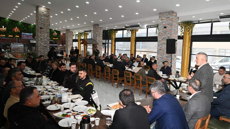 Geniş bir salonda masalarda oturan kalabalık topluluk ve elinde mikrofonla konuşan bir kişi görülüyor.